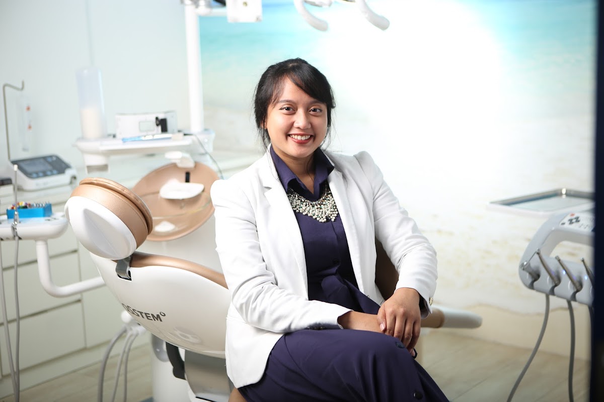 A smiling female dentist in a white coat seated confidently next to a modern dental chair in a clean, well-lit treatment room.