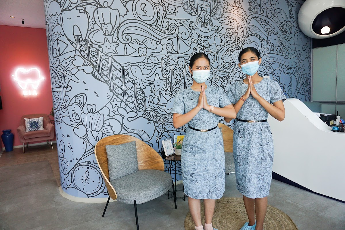 Two reception staff in matching uniforms greet with a traditional Balinese namaste pose in a stylish lobby featuring artistic mural walls and a neon tooth sign.