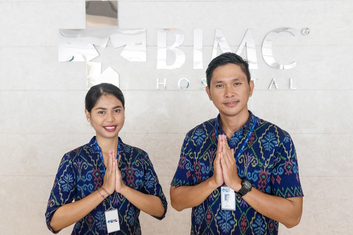 Two BIMC Hospital staff in batik uniforms offering welcoming gesture in front of clinic signage