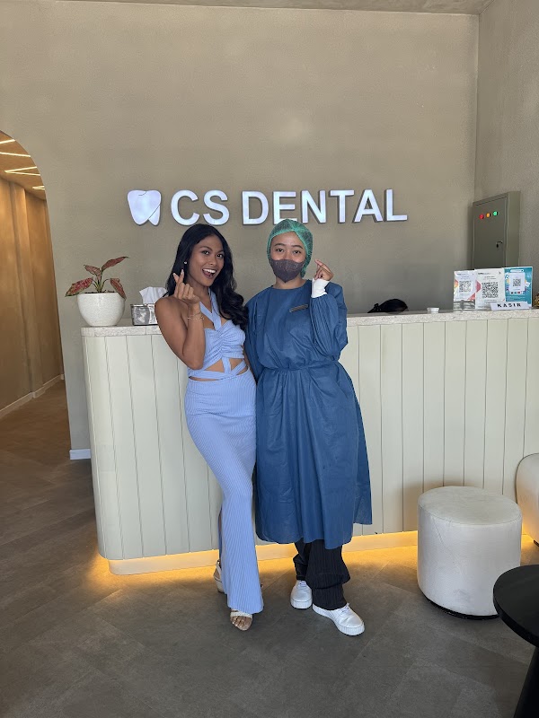 A dental professional in blue scrubs and mask poses with two happy international clients at the CS Dental reception desk, showcasing the clinic's welcoming atmosphere.