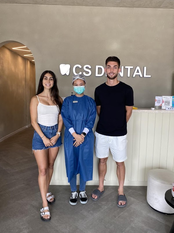 A masked dental staff member in blue scrubs poses with a smiling client in front of the well-lit CS Dental reception counter with fresh tulips visible.
