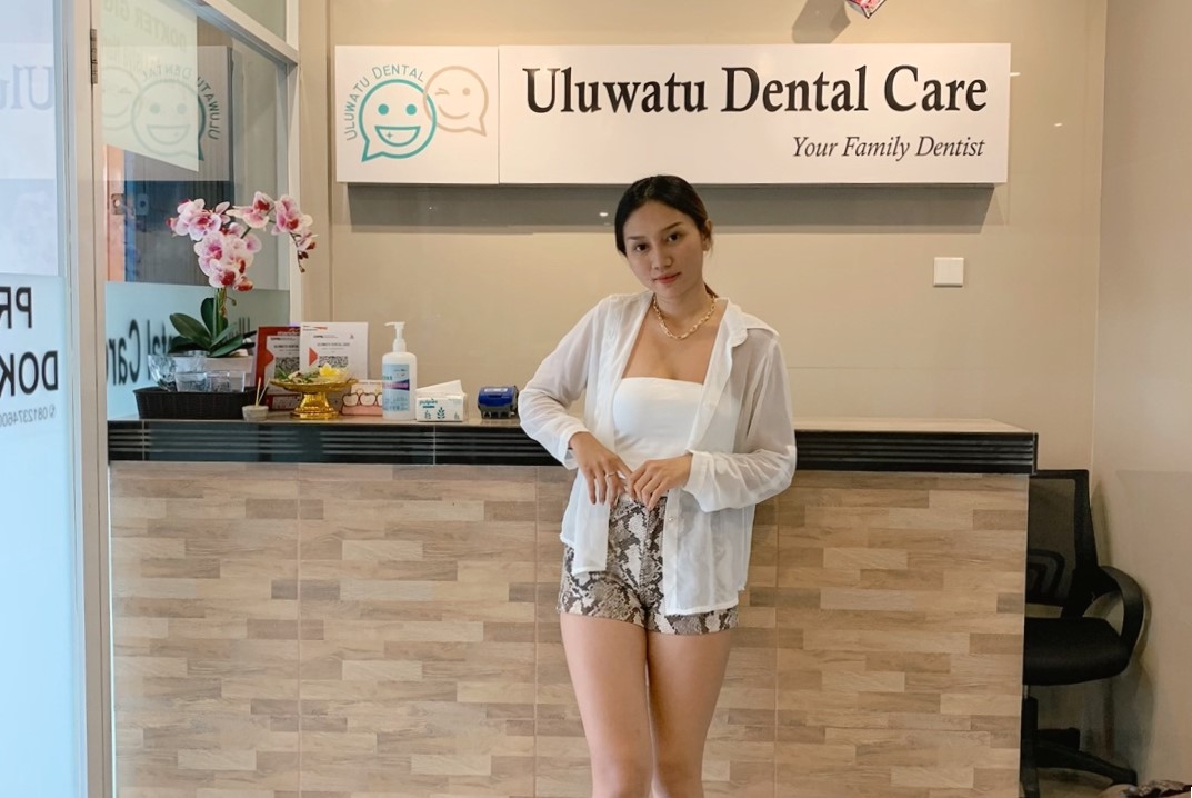 A client poses in front of the Uluwatu Dental Care reception desk, showcasing a clean and tidy interior with flowers and branded signage.