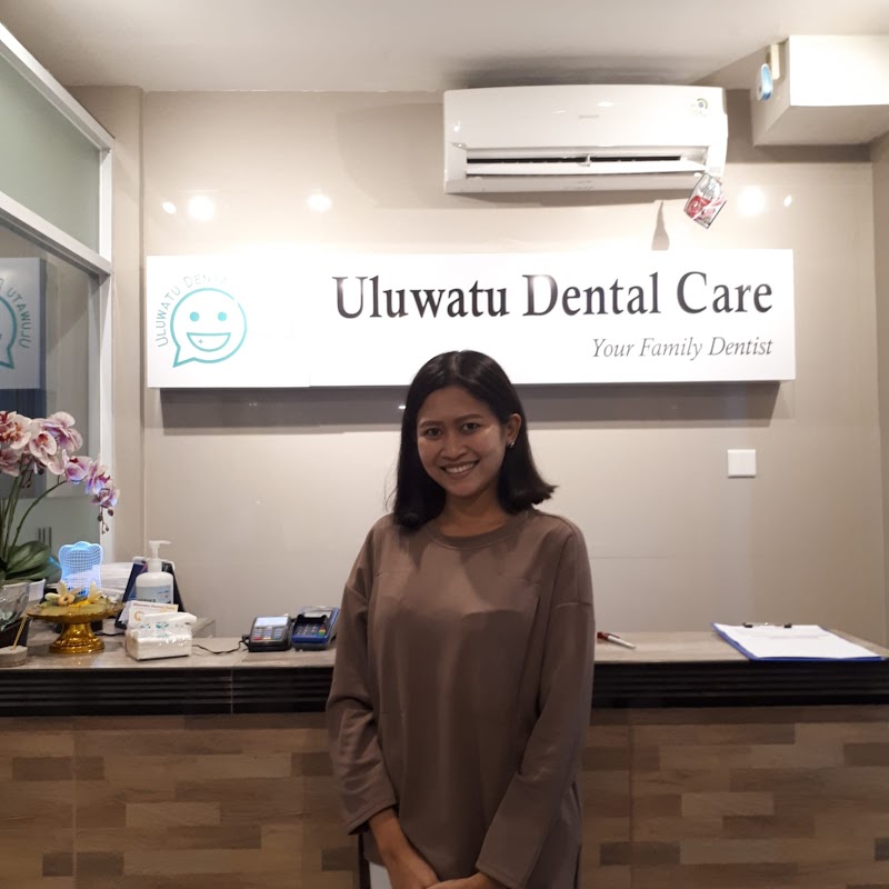 A friendly receptionist smiles in front of the 'Uluwatu Dental Care' sign at the front desk area, though the lighting is somewhat dim and exposed AC unit and miscellaneous items detract slightly.